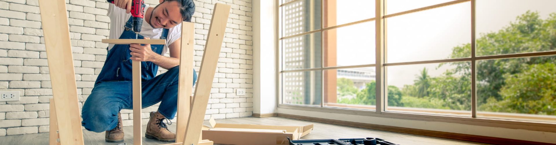 A handyman is working hard on a wooden frame in front of a window.